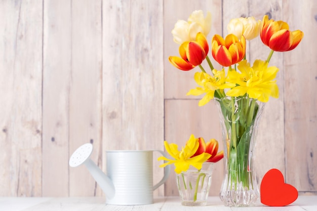 Tulip flower arrangement in glass vase with heart greeting watering can decor on wooden table background wall close up Mother's Day design concept