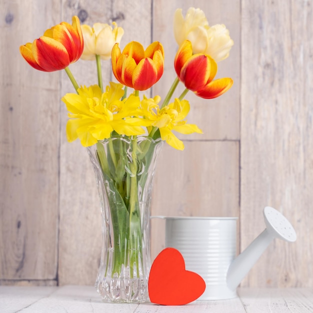 Tulip flower arrangement in glass vase with heart greeting watering can decor on wooden table background wall close up Mother's Day design concept