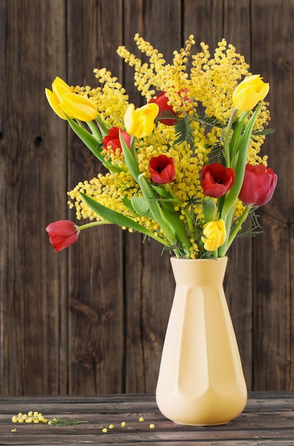 Tulips and mimosa in yellow vase on dark wooden background