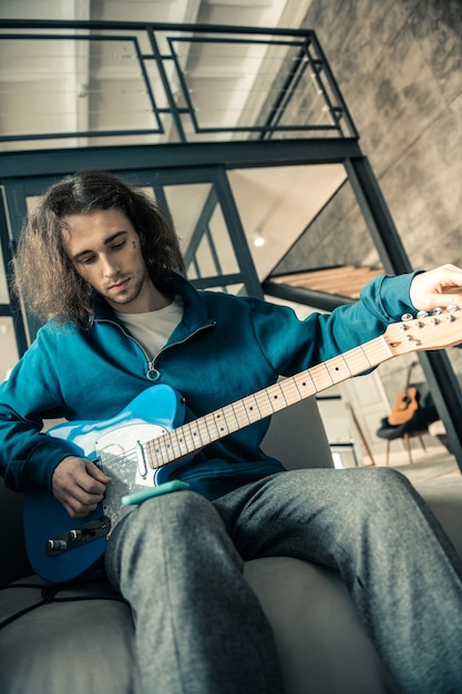 Tuning guitar. Dark-haired attentive man wearing blue sweatshirt and preparing guitar for daily repetition at home