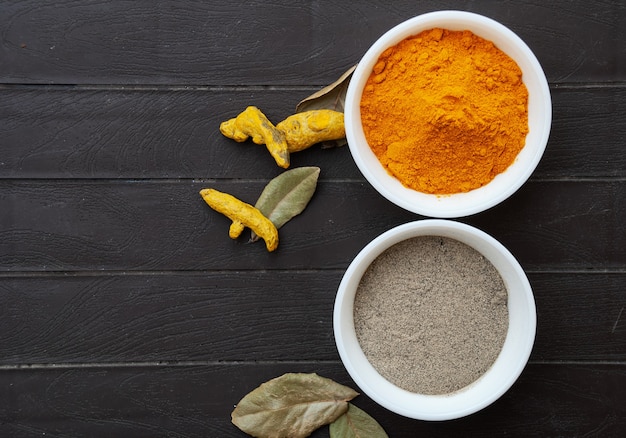 Turmeric powder and black pepper powder in white bowls, turmeric roots and bay leaves next to the bowls