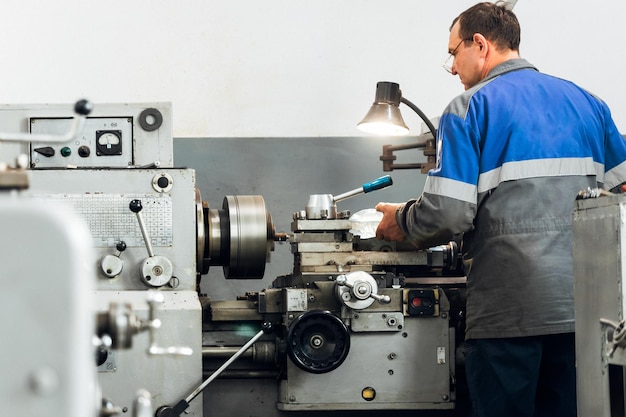 Turner stands behind lathe in production hall and works View of worker from behind from back in overalls