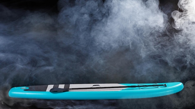 Turquoise inflatable SUP board with a paddle on a black background in the fog