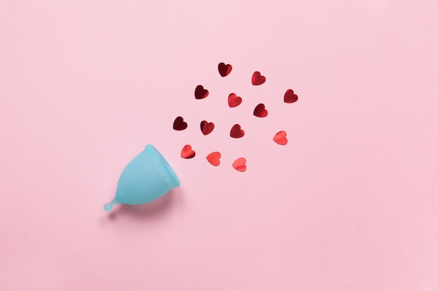 Turquoise menstrual cup on pastel pink background with red hearts confetti. Feminine hygiene product