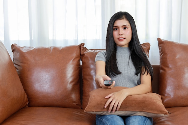 TV and happiness concept. Beautiful asian woman in casual sitting on sofa in living room, holding television remote and watching something with happy smiling face. Lifestyle concept.