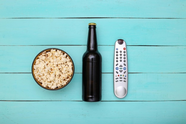 TV time. Beer bottle, tv remote and popcorn bowl on blue wooden background. Top view