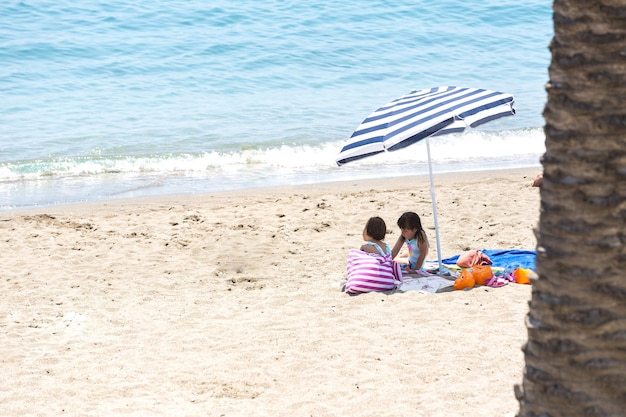 Twin sisters sitting on towels on the beach under the shade of an umbrella with a bag and armbands around them Concept of beach vacation sun protection sea lifestyle and relaxation