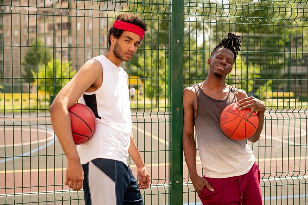 Two active guys in sportswear standing by fence surrounding basketball court while having rest after training