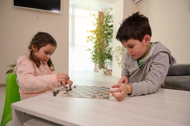 Two adorable Caucasian elementary aged kids, boy and girl, brother and sister having great time playing checkers board game together at home interior.