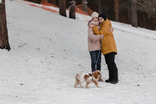 Two adorable young girls having fun together by beautiful frozen park forest Cute sisters playing in a snow with a dog