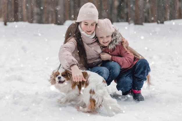 Two adorable young girls having fun together by beautiful frozen park forest Cute sisters playing in a snow with a dog