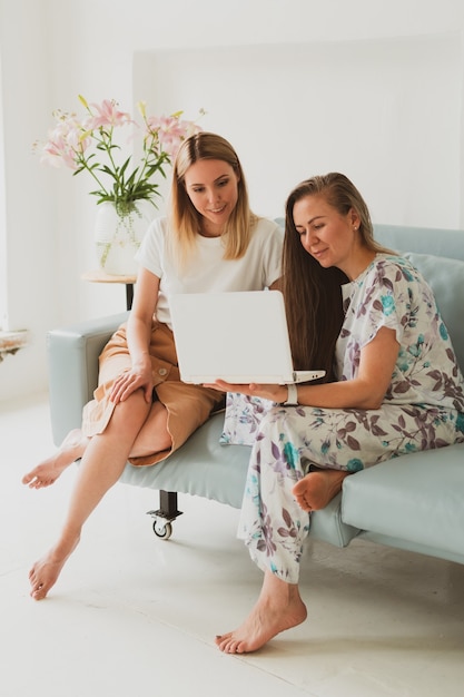 Two adorable young women chatting at home on the couch, drinking coffee and working on a laptop