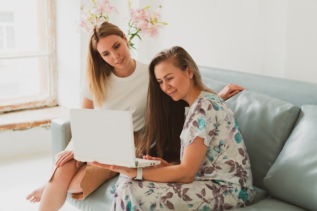 Two adorable young women chatting at home on the couch, drinking coffee and working on a laptop