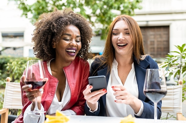 Two adult woman having fun during happy hour after work colleagues laughing while looking at smart phone social network and mobile app for tech addicted people