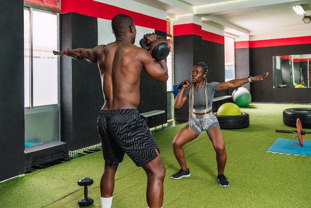 Two African bodybuilder athletes doing toning exercises for biceps and shoulders with kettlebells in a gym. African bodybuilders doing crossfit workout routine