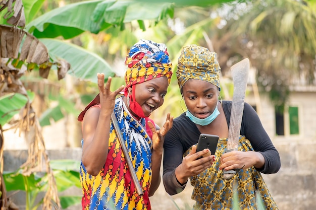 two african female farmers with nose mask and farming tools looking happily into a smartphone