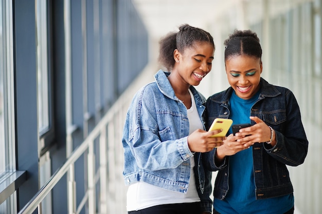 Two african woman friends in jeans jacket using mobile phones indoor together.