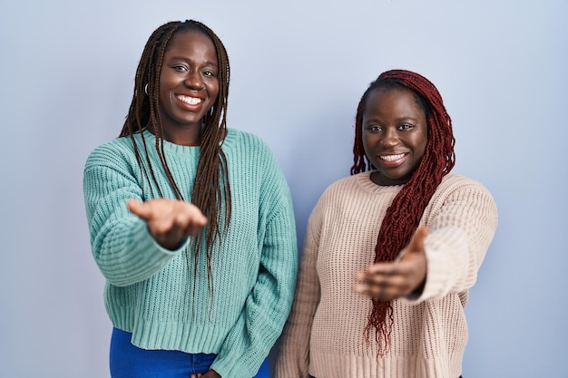 Two african woman standing over blue background smiling cheerful offering palm hand giving assistance and acceptance.