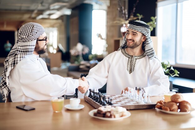 Two arab businessmen shake hands behind chess board.