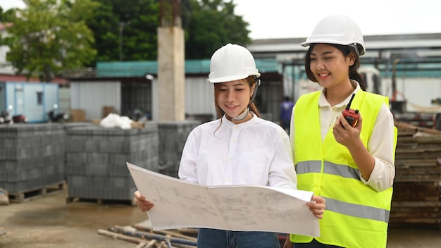 Two Architect women wearing safety helmet and yellow vests inspecting industrial building construction site