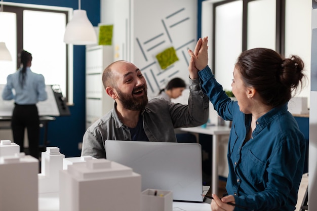 Two architects high fiving sitting at desk in architectural modern office with laptop and foam scale model of residential buildings. Successful happy smiling colleagues celebrating team project.