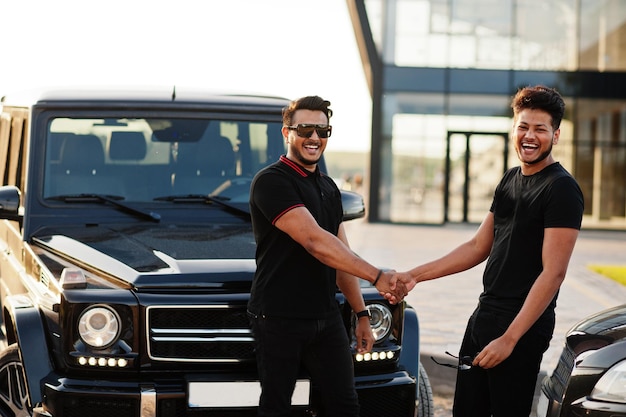 Two asian brothers man wear on all black posed near suv cars and hand shaking.