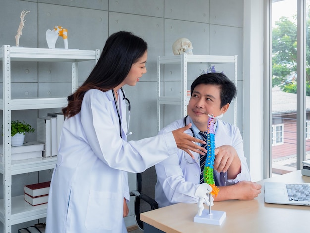 Two Asian doctors in white uniforms talking and working together in medical office The orthopedic adult male doctor provides knowledge and counseling to female doctor about spine on desk in hospital