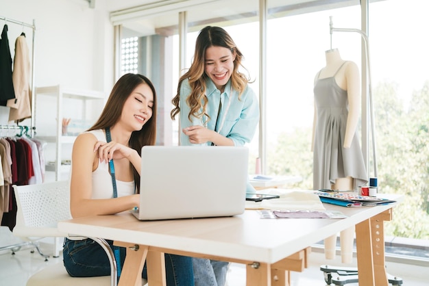 Two Asian female fashion designers or dressmakers brainstormed and discuss the design of the new collection with examples of fabrics and sketches on the laptop Working together in the studio