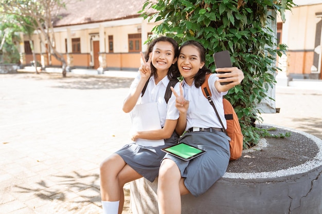 Two asian high school girls selfie with v hand gesture using cell phones in school park