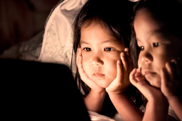 Two asian little girls using digital tablet under blanket at night in vintage color tone