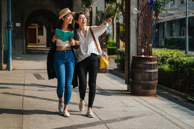 two asian tourists sisters travel in little village in europe. happy girls travelers having fun in creative old town with wind barrel in street. women holding guide book and point view with sun flare