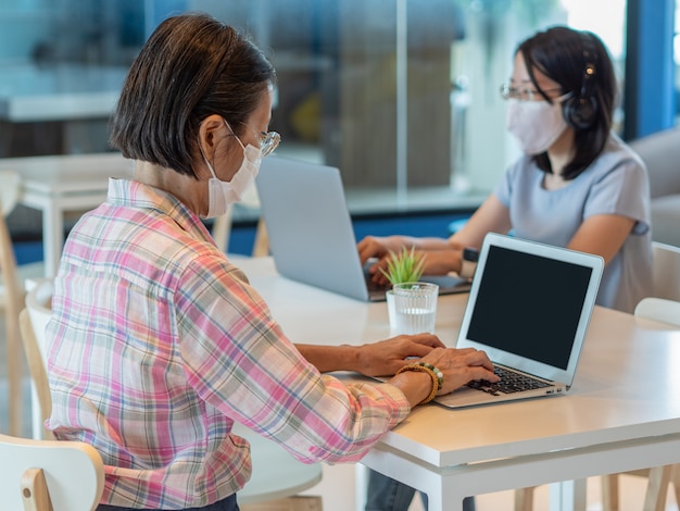 Two asian women wearing face mask and using smartphone and laptop for video calling or working, sit on separate tables for keeping safety social distancing, as new normal lifestyle concept.