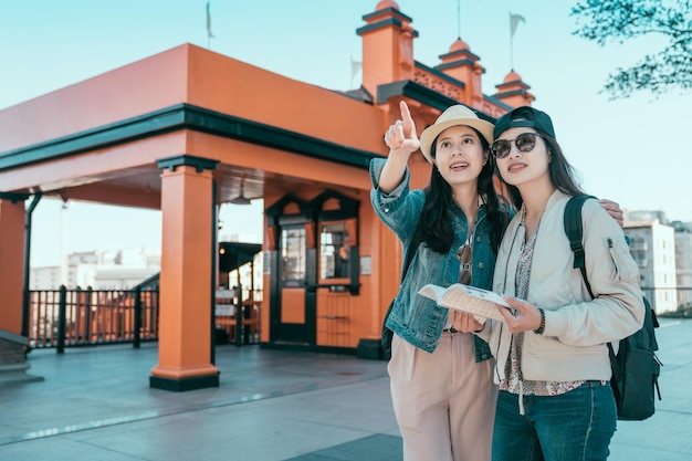 two asian young girl friends standing outdoor travel in graduation trip on summer. beautiful sisters backpackers holding guide book searching direction point front. angels flights railway station.