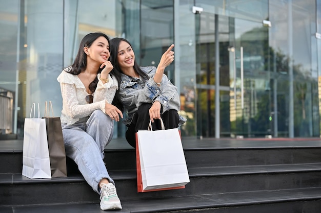 Two attractive Asian sitting on the stairs in front of the mall entrance with their shopping bags