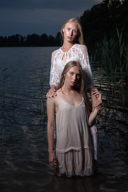 Two attractive caucasian twin sisters with long blonde hair posing in light dresses in water of lake at summer night