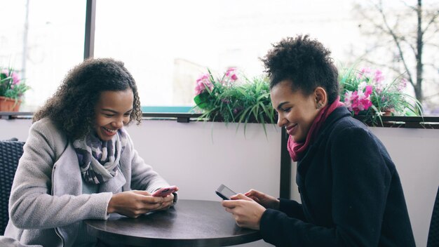 Two attractive mixed race female friends sharing together using smartphone in street cafe outdoors