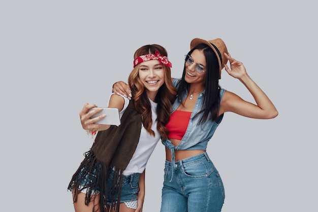 Two attractive stylish young women taking selfie and smiling while standing against grey background