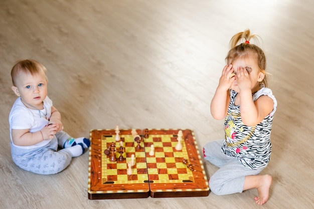 Two babies boy and girl playing chess on the white wooden floor at home.