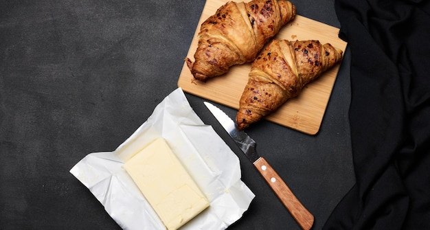 Two baked croissants a pack of butter and a knife on a black background top view