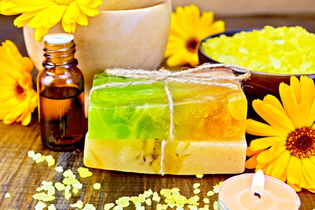 Two bars of homemade soap, yellow bath salt, candle, oil in a bottle and marigold flowers on a background of wooden planks