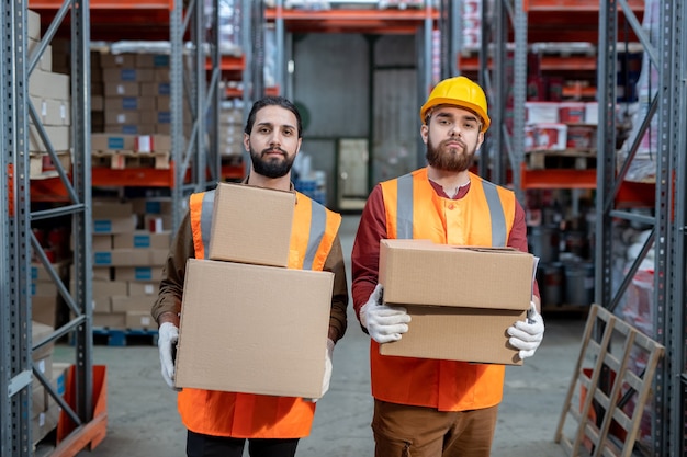 Two bearded workers of warehouse holding boxes while moving along aisle