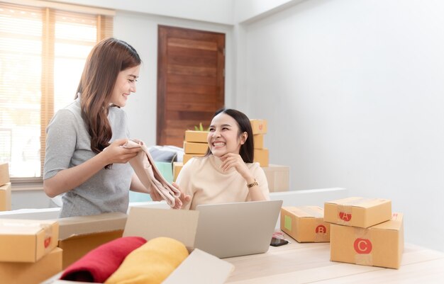 Two beautiful Asian women are checking orders by laptops via the internet. And pack the paper box With a happy smiling face, being a new normal online business