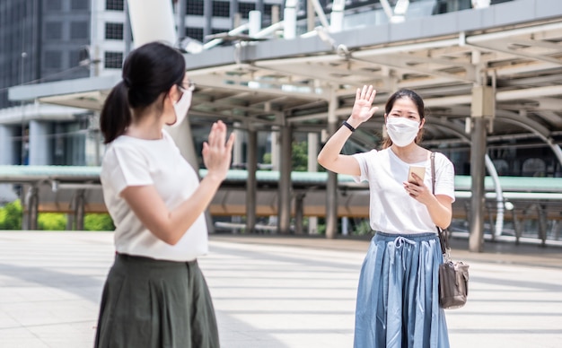 Two beautiful Asian women greeting with colleague, wearing a disposable medical face mask every time outside the house, as a new normal trend and self-protection against Covid19 infection.