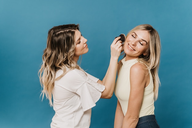 Two beautiful blondes wearing casual clothes help each other make up before going to a party, standing on a blue background the concept of friendship and beauty.