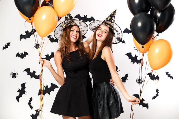 Two beautiful brunette girls dressed in black dresses and witch hats hold black and orange balloons. Halloween .