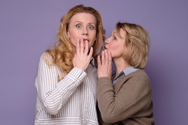 Two beautiful caucasian mature women telling secret on a colored background