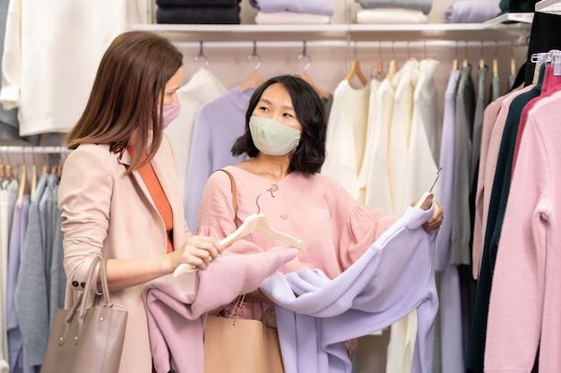 Two beautiful friends in masks choosing jumpers and dresses together during their shopping in clothing store