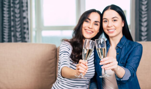 Two beautiful girlfriends in casual clothes rest sitting on the couch talking and drinking champagne