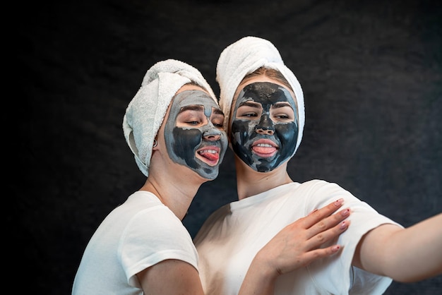 Two beautiful girls applying clay black mask isolated on black Home take care of face skin Lifestyle