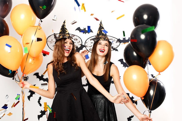 Two beautiful girls in black dresses and witch hats have fun with black and orange balloons and confetti.Halloween party .